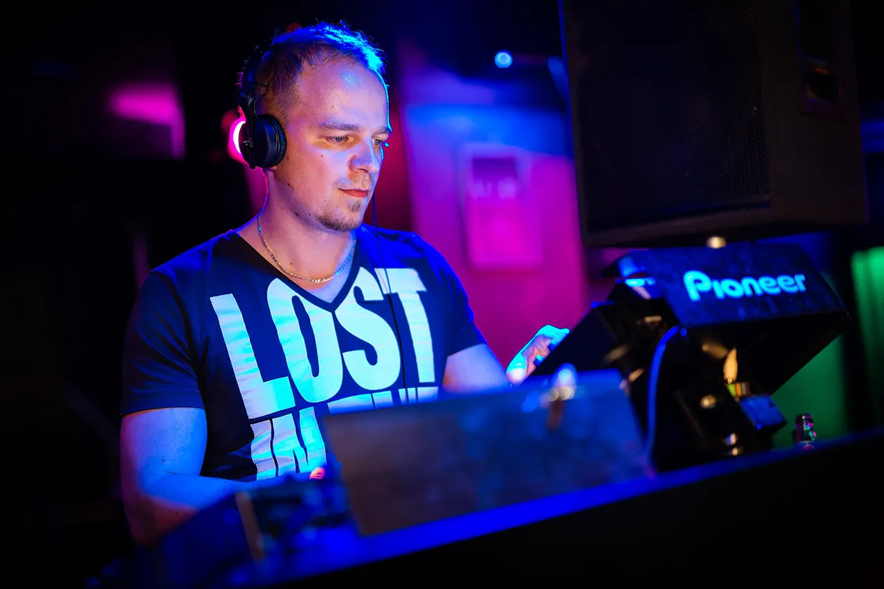 Muž, DJ Acidflash, stojící za přehrávači a mixující taneční hudbu na party v barevně nasvíceném klubu
