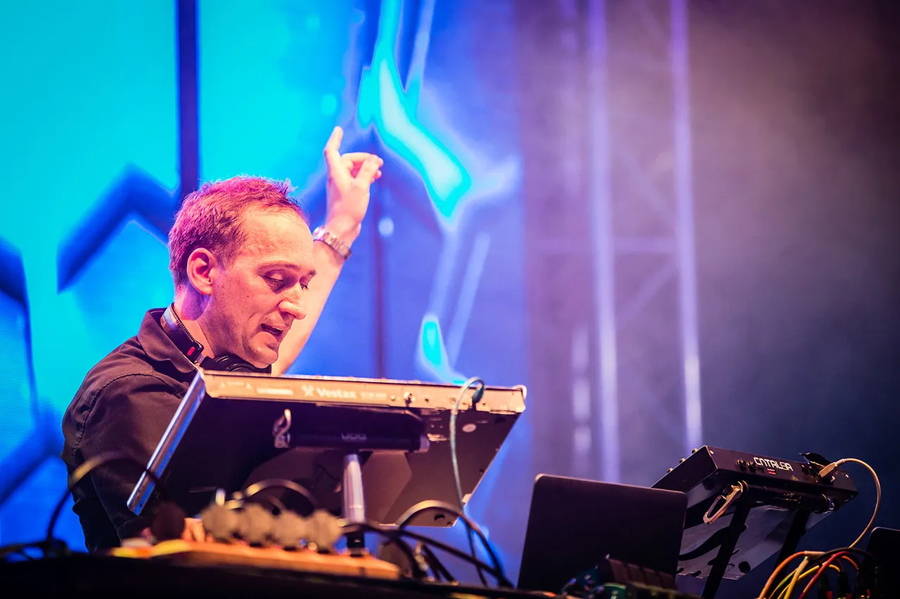 Muž, Paul van Dyk, stojící za mixážním pultem, povzbuzující publikum levou rukou zdviženou vzhůru v barevném industriálním prostředí halové taneční party