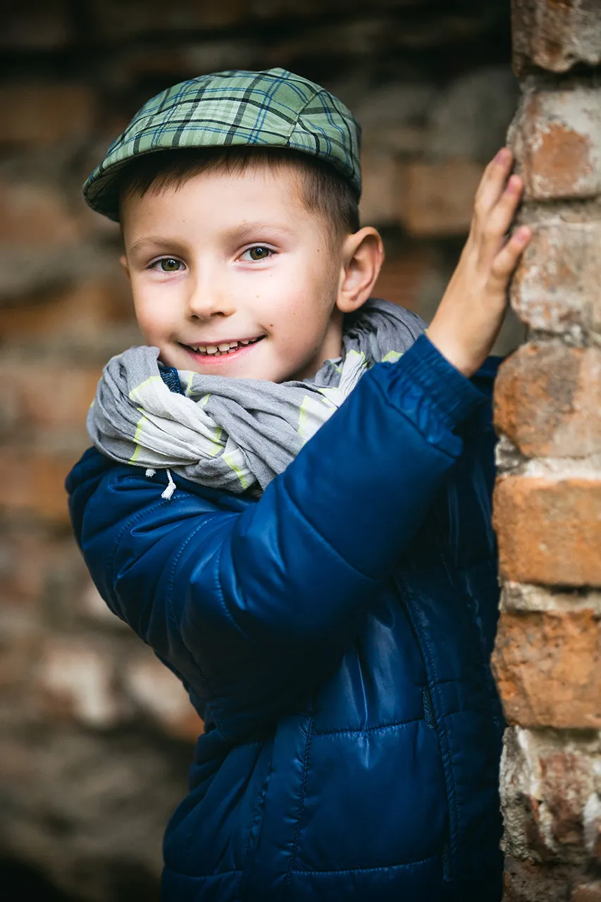 Usmívající se mladý chlapec s zelenou čepicí bekovkou, v modré bundě s šedou šálou kolem krku, opřený o cihlovou zeď.