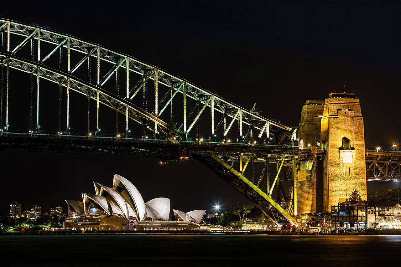 Noční pohled na osvětlenou operu v Sydney a část mostu Harbour Bridge přes záliv.